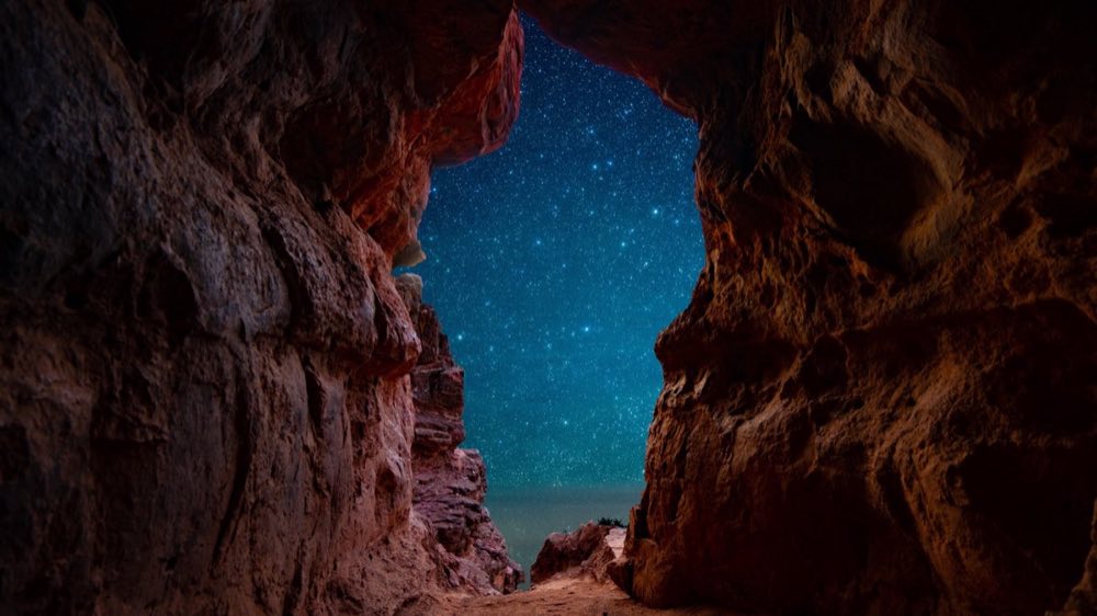 A star-filled night sky seen through the opening of a rocky cave